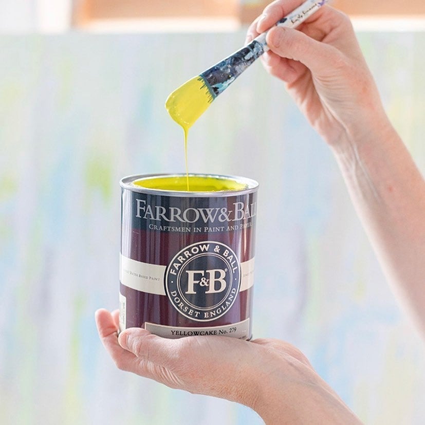 Person holding a Farrow & Ball paint can with a brush dripping yellow paint.