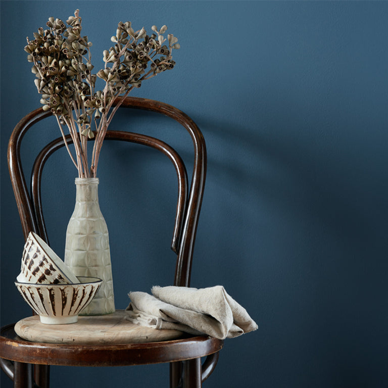 Decorative still life with a vase, bowl, and dried flowers on a wooden chair against a dark blue wall.