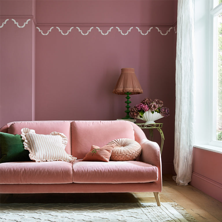 Pink sofa in a living room with a lamp and flowers on a table against a pink wall.