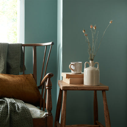 Wooden chair with a cushion next to a wooden side table with a vase and books against a green wall.