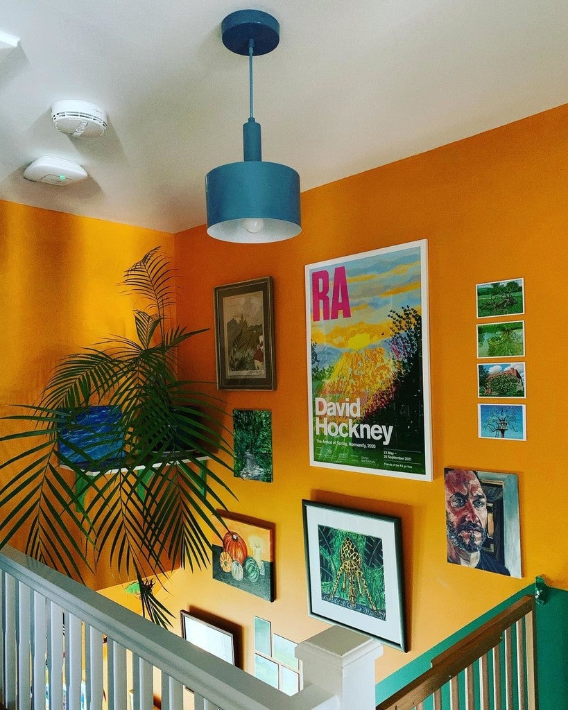 Stairwell with orange walls, a blue pendant light, and various framed artworks including a David Hockney poster.
