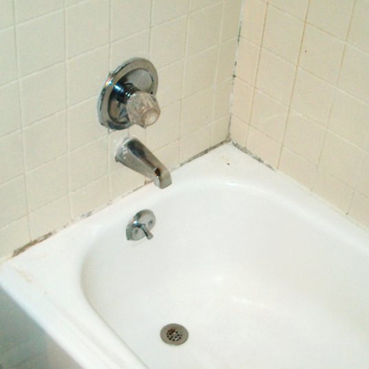 Corner of a white bathtub with chrome faucet and handles against a tiled wall.