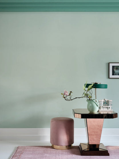 Pink ottoman and table against a light green wall with a lamp and plant on the table.