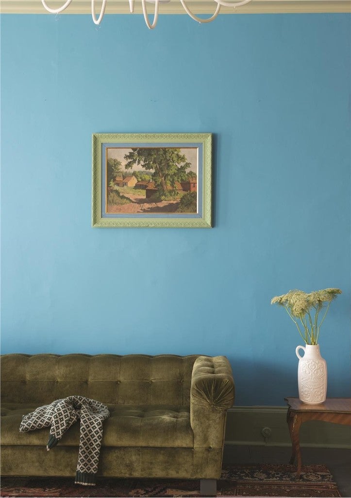 Green sofa with a patterned throw, framed painting on a blue wall, and a vase with flowers on a small table.