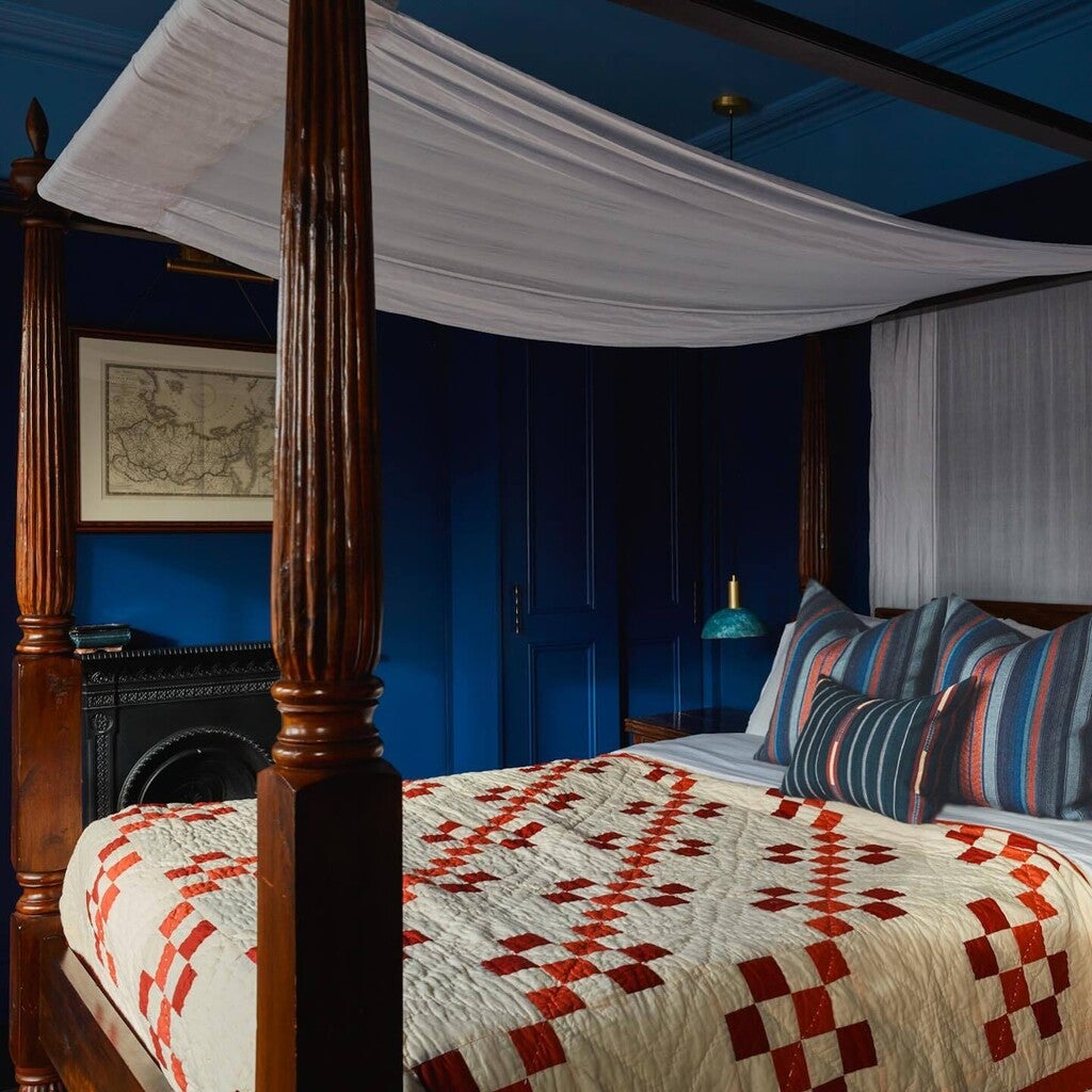Bedroom with a four-poster bed featuring a patterned quilt and striped pillows against a blue wall.