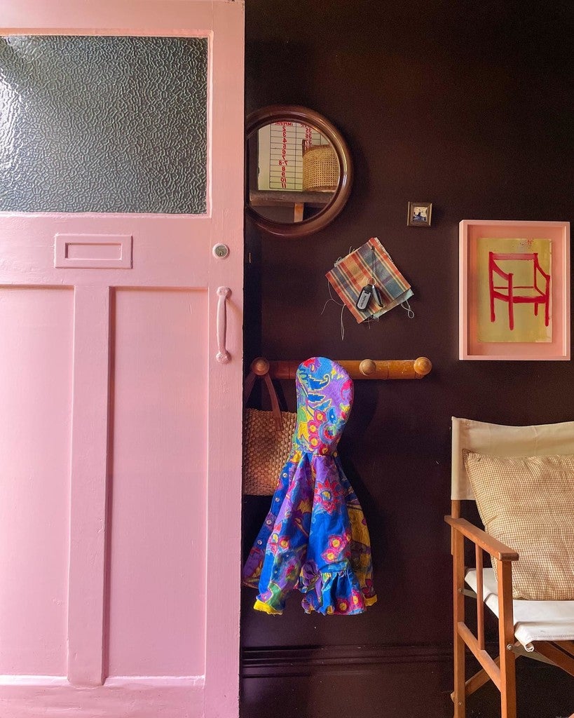 Pink door with colorful raincoat, mirror, and framed picture on a dark wall.