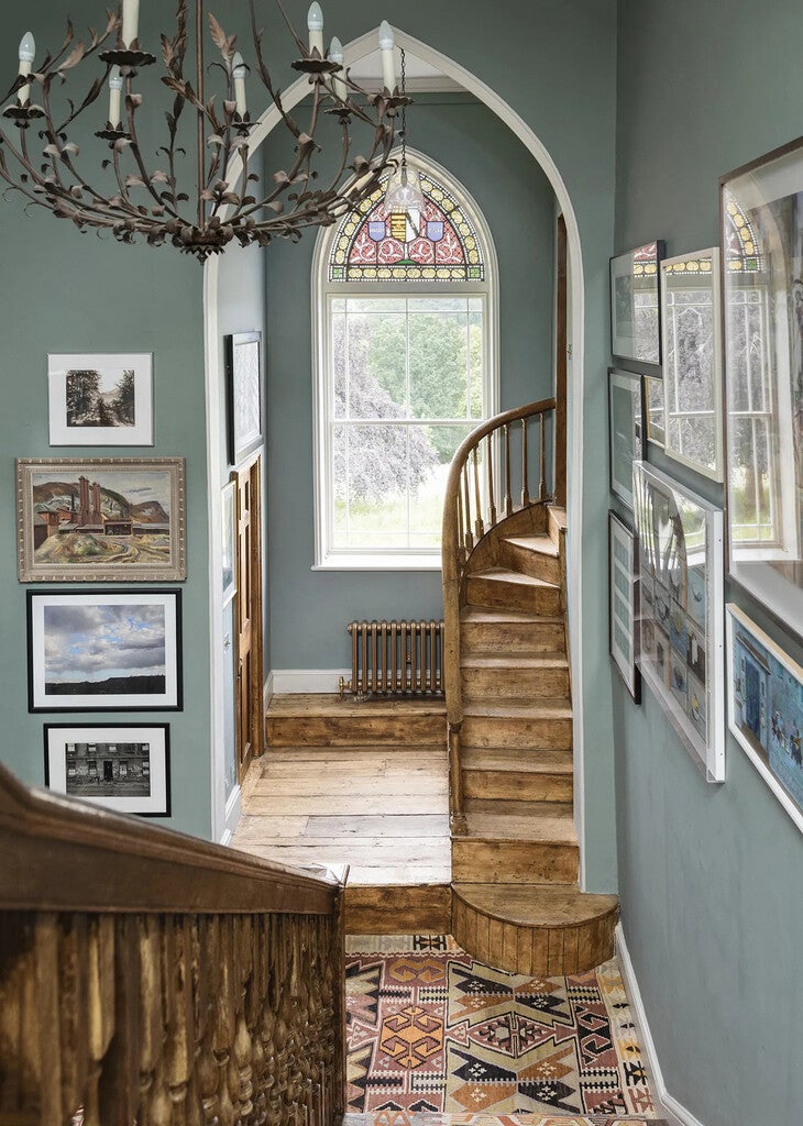 Staircase with wooden banister and chandelier in a room with stained glass window and framed pictures.