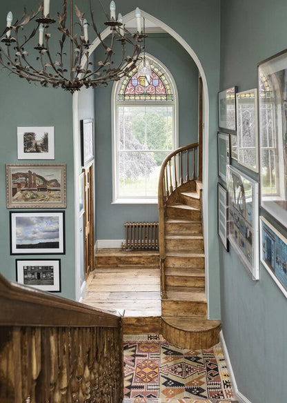 Staircase with wooden banister and chandelier in a room with stained glass window and framed pictures.