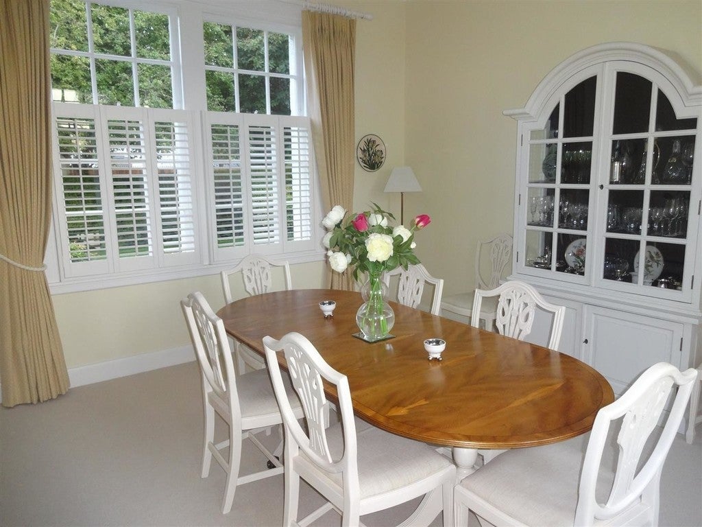 Dining room with wooden table and white chairs, large window with shutters, and decorative cabinet.