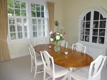 Dining room with wooden table and white chairs, large window with shutters, and decorative cabinet.