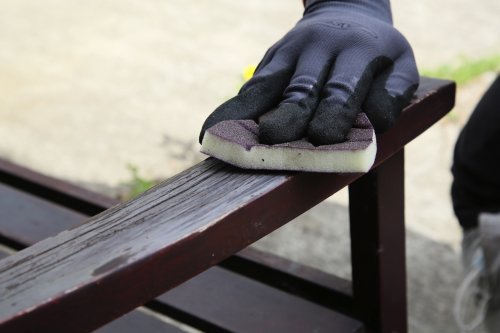 Person cleaning a wooden bench with a sponge and black gloves.