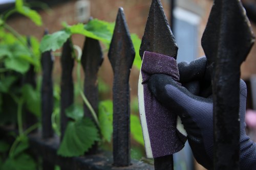 Person wearing a glove holding a purple sanding block against a metal fence.