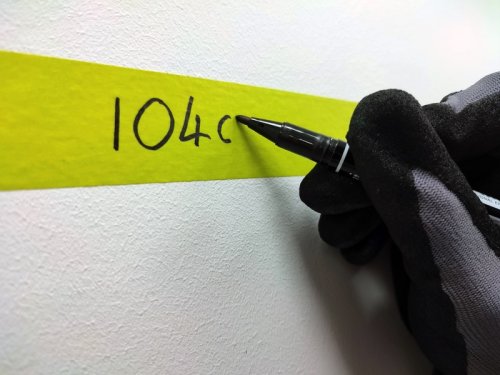 Hand wearing a black glove writing '104c' on a yellow strip of tape with a pen.