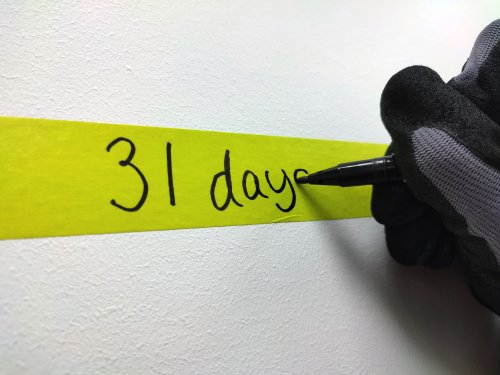 Hand wearing a black glove writing '31 days' on a yellow strip of paper with a pen.