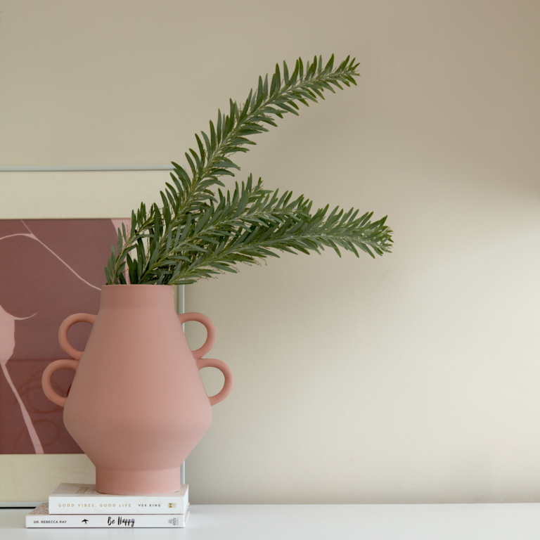Pink vase with green plant on a white surface against a beige wall