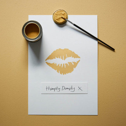 Gold paint can, brush, and paper with a gold lip print and 'Humpty Dumpty X' text on a beige background.