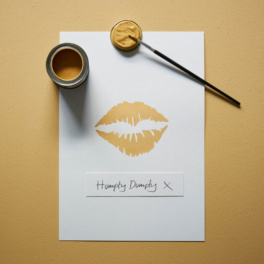 Gold paint can, brush, and paper with a gold lip print and 'Humpty Dumpty X' text on a beige background.