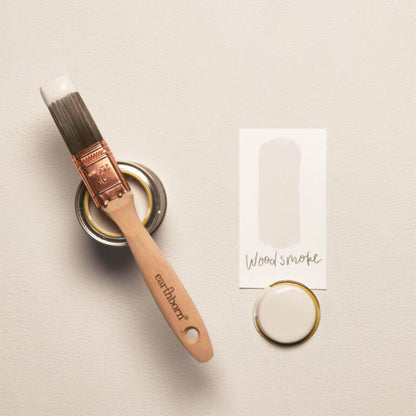 Paintbrush with 'earthborn' branding next to a small container of paint and a color swatch labeled 'Wood smoke' on a beige background.