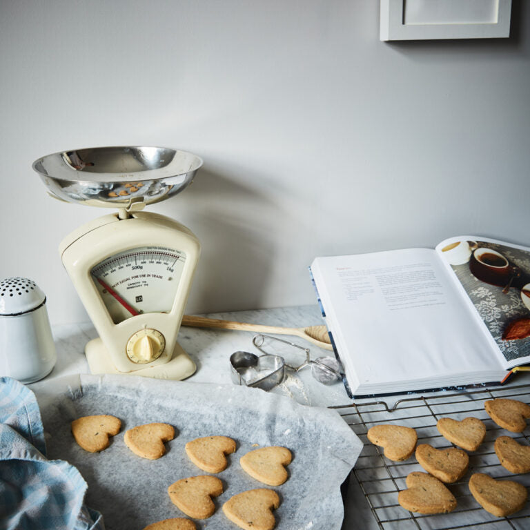 Baking scene with heart-shaped cookies, a scale, and a cookbook on a table.