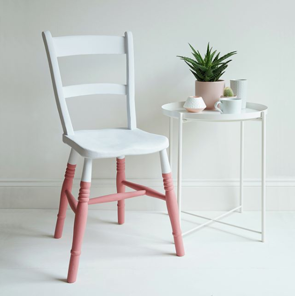 White chair with pink legs next to a small round table with a plant and cups on a light background