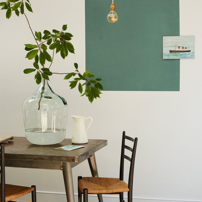 Dining area with a wooden table, chairs, a vase with greenery, and a painting on the wall.