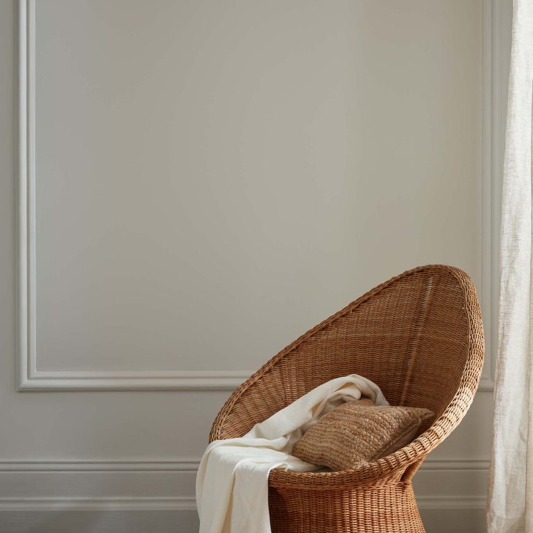 Wicker chair with a white blanket and brown pillow against a plain wall.