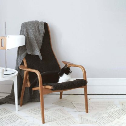 Wooden armchair with a black cushion and gray blanket, featuring a cat, in a minimalistic room.