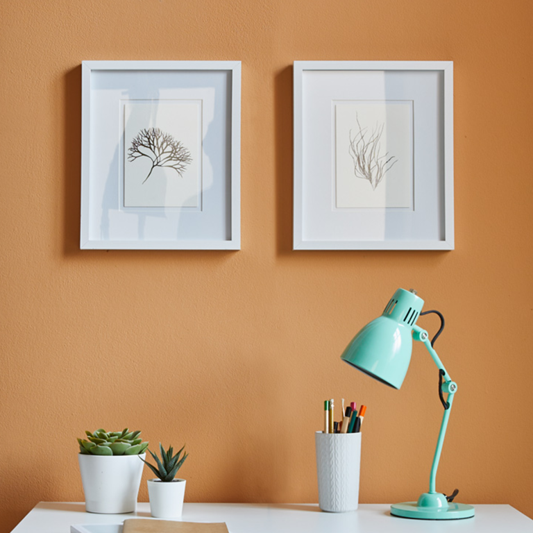Two framed artworks on an orange wall with a desk featuring a lamp, plants, and stationery.