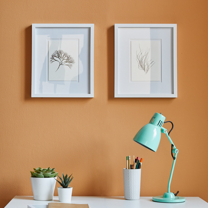 Two framed artworks on an orange wall with a desk featuring a lamp, plants, and stationery.