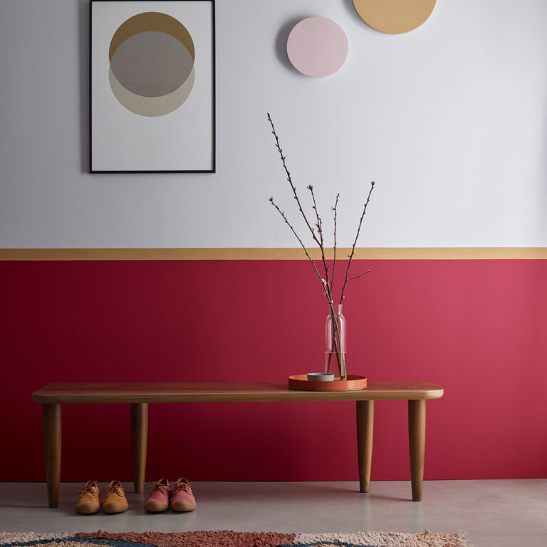 Wooden bench against a wall with a red stripe, a vase with branches on a small table, and abstract art on the wall.