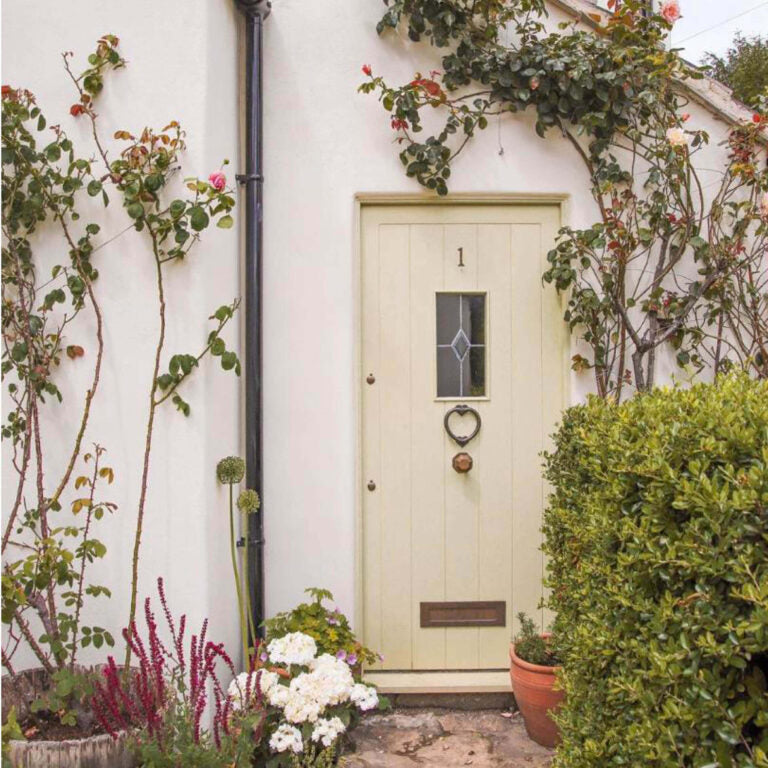 Cottage door with decorative heart-shaped handle, surrounded by plants and flowers.