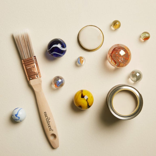 Paintbrush with 'earthborn' branding next to marbles and a paint can on a beige background