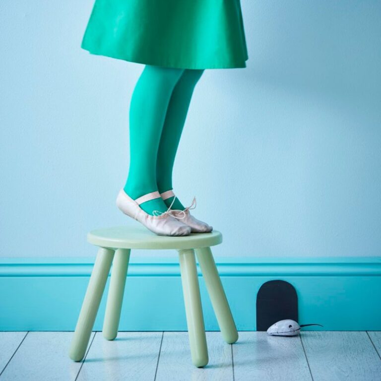 Person wearing a green skirt and leggings standing on a small stool against a light blue wall.