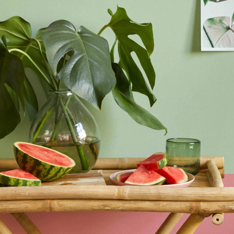 Sliced watermelon on a wooden tray with a green plant and glass in the background.
