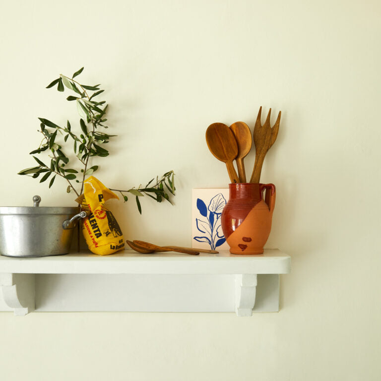 White shelf with kitchen items including a pot, plant, and wooden utensils against a light wall.