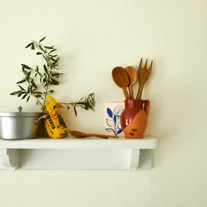 White shelf with kitchen items including a pot, plant, and wooden utensils against a light wall.