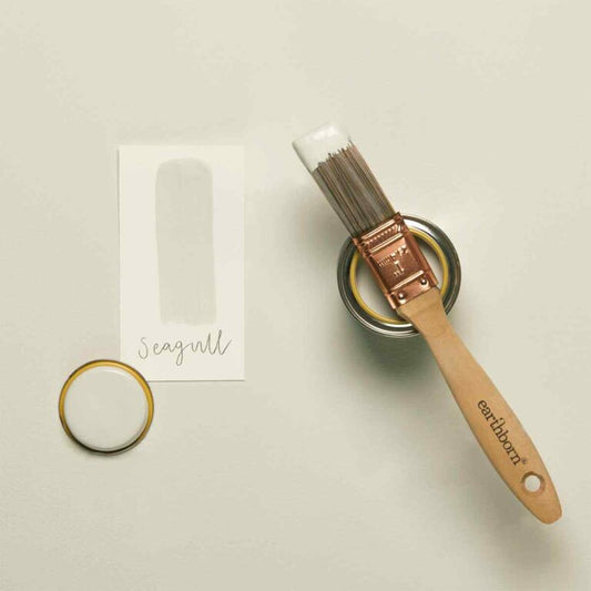 Paintbrush with wooden handle and metal ferrule, next to a small container of paint and a ring on a light background.