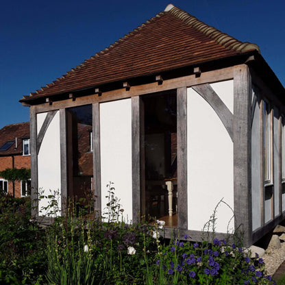 Wooden gazebo with a white wall and brown roof in a garden setting.
