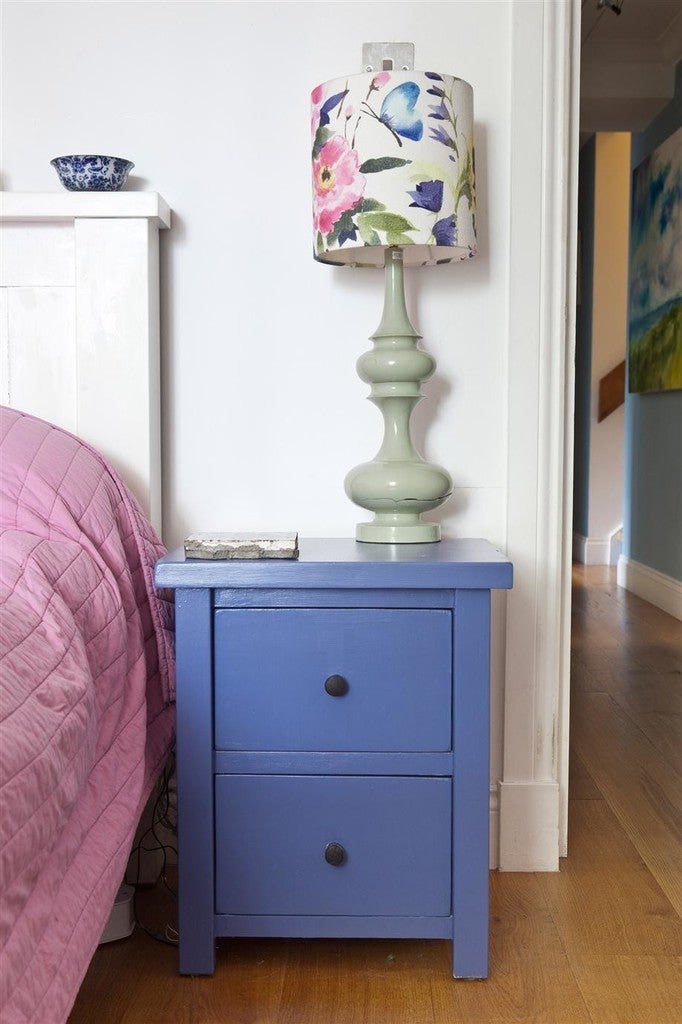 Blue nightstand with a green lamp featuring a floral shade next to a bed with pink bedding.