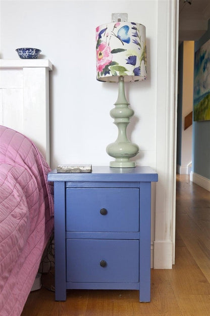 Blue nightstand with a green lamp featuring a floral shade next to a bed with pink bedding.