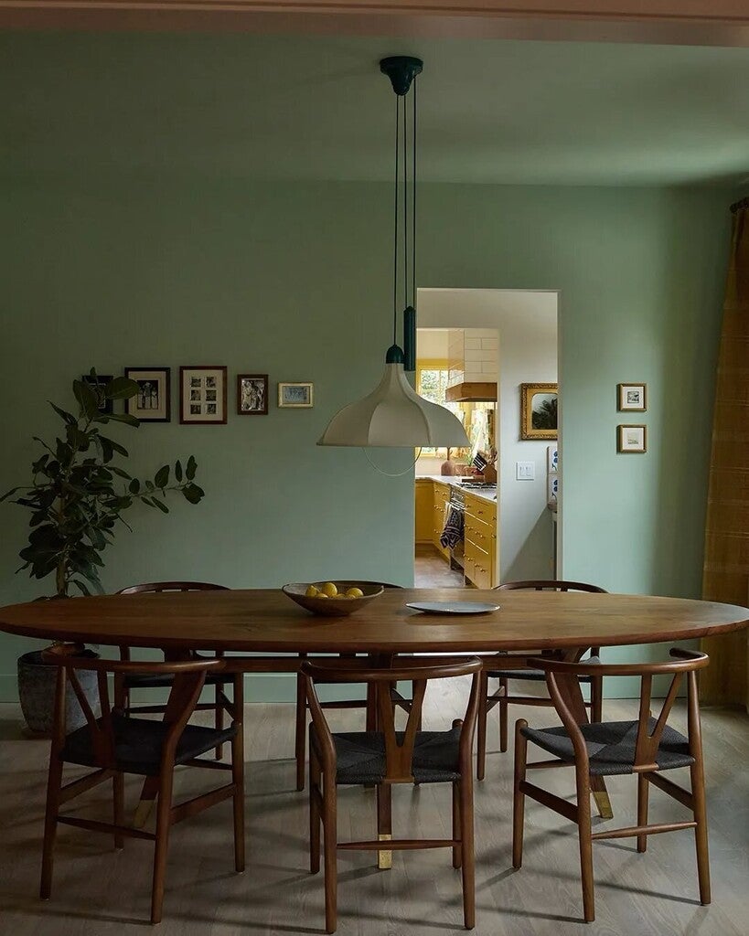 Dining area with wooden table and chairs in a room with green walls and a kitchen in the background.