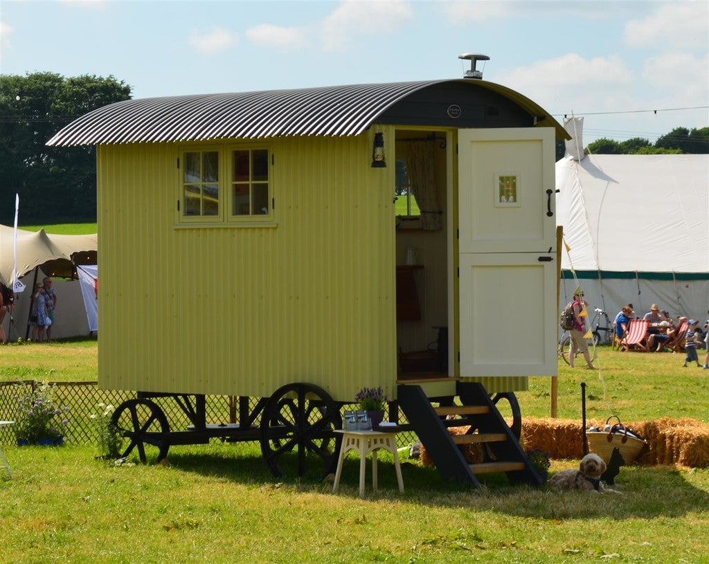 Yellow shepherd's hut with an open door in a grassy field with people and tents in the background.