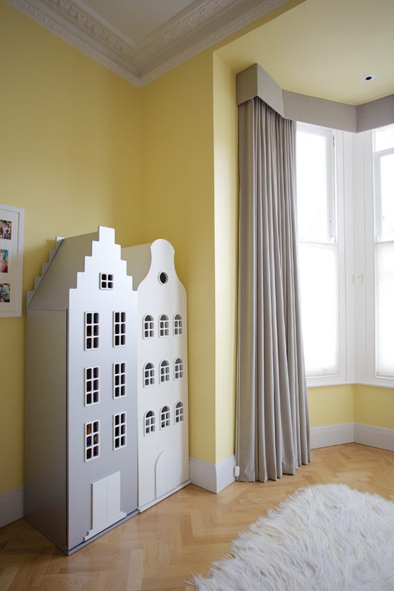 White children's playhouse against a yellow wall with a large window and curtains.