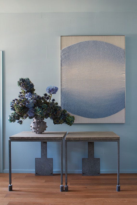 Two tables with decorative items against a light blue wall with a piece of abstract art.